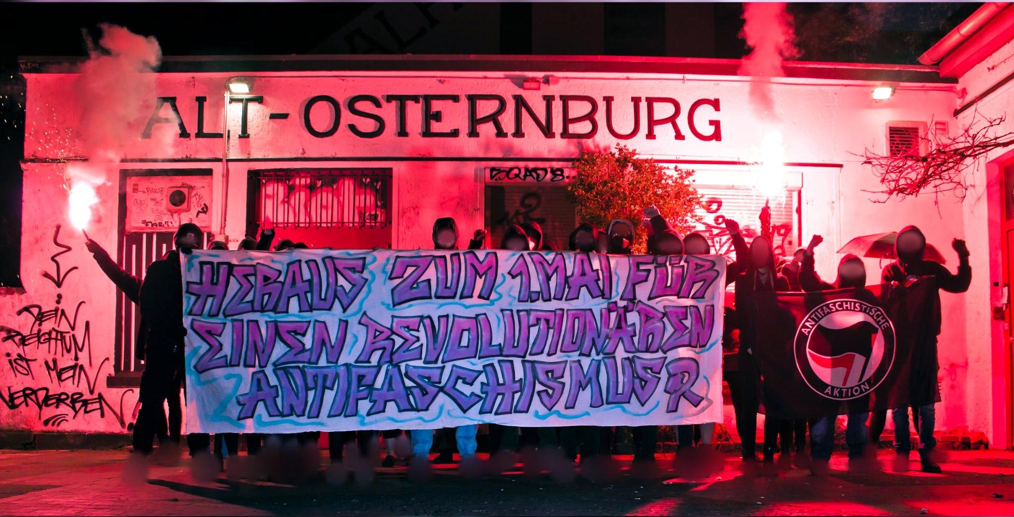 Das Foto bildet eine große Gruppe schwarz vermummter Menschen ab, die ein Transparent und eine Antifa-Fahne halten. Links und rechts brenen Menschen rote Pyro-Fackeln ab. Auf dem Transpi steht: "Heraus zum 1. Mai - Für einen revolutionären Antifaschismus!"